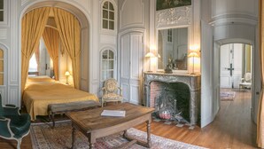 A room with a bed, a chair, a table, a fireplace, and a mirror.