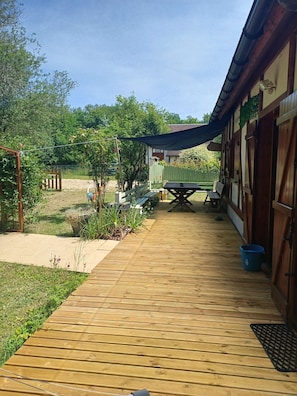 Eine Holzterrasse führt zu einem Gebäude mit überdachtem Bereich, einem grünen Zaun und einer Bank.