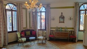 Ein Zimmer mit einer Couch, zwei Stühlen, einem kleinen Tisch, einer Kandelaber und Blick durch große Fenster.