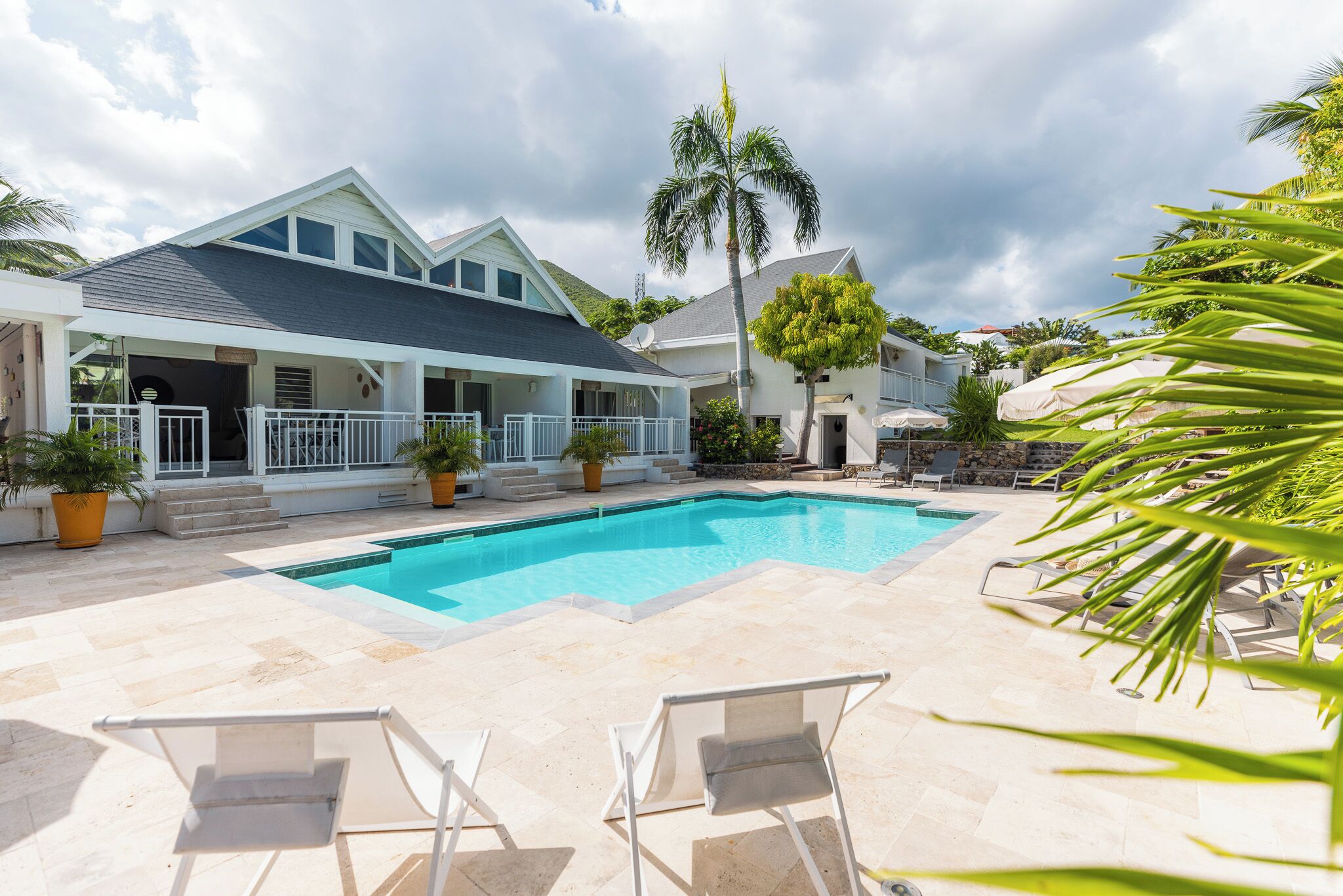 A swimming pool with lounge chairs, a covered patio area, and a white house with a dark roof.