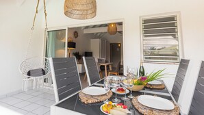 En matplats med ett bord dukat för fyra personer, inklusive tallrikar, glas och en flaska champagne. Ovanför bordet hänger en lampa av flätat material, och det finns en vit stol med ett flätat säte. Rummet har en modern design med ett stort fönster och en dörr som leder till ett annat område.