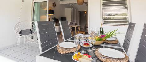 En matplats med ett bord dukat för fyra personer, inklusive tallrikar, glas och en flaska champagne. Ovanför bordet hänger en lampa av flätat material, och det finns en vit stol med ett flätat säte. Rummet har en modern design med ett stort fönster och en dörr som leder till ett annat område.