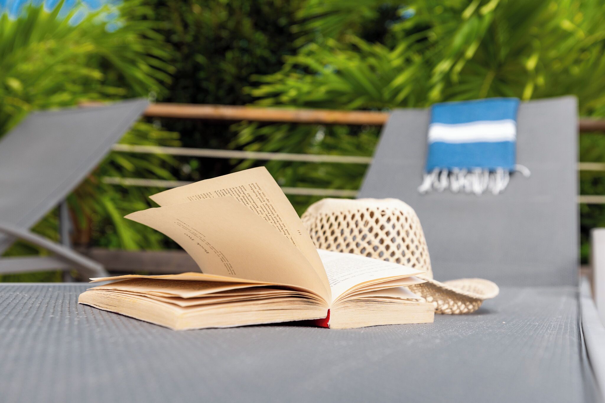 Un livre ouvert posé sur une chaise longue, avec un chapeau tressé à côté, le tout sur fond de verdure.