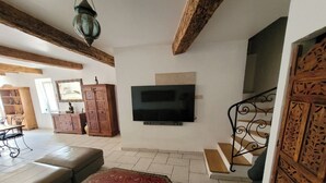 A living room with a TV, wooden beams, a sofa, a patterned ottoman, a cabinet, and a staircase. - In a quiet location between Aix and Marseille. Provence. Beach 10 km away, with no buildings directly across from the property  (Lançon-Provence)