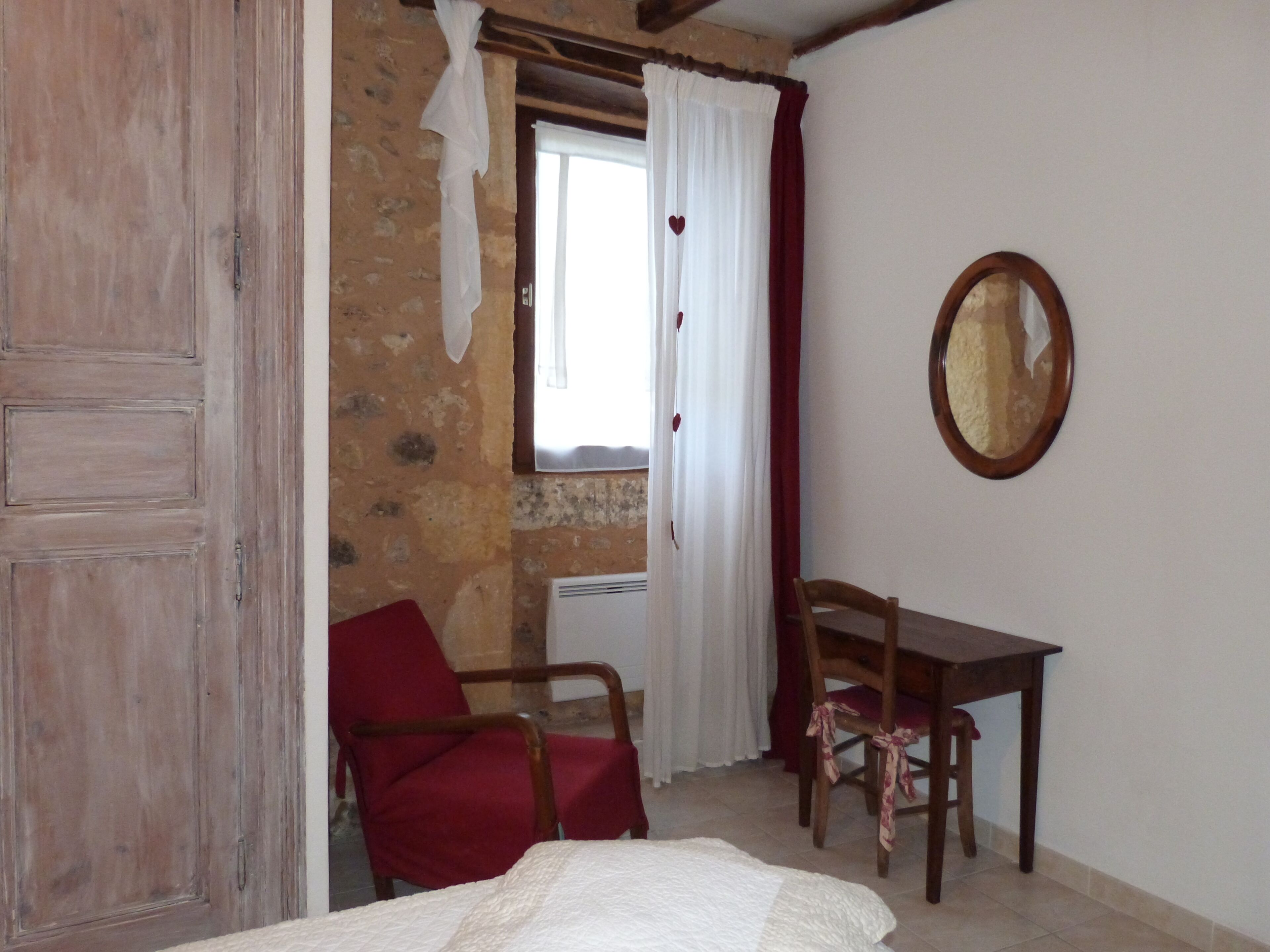 A room with a wooden door, a red chair, a wooden desk, a white bed, a window with white curtains, and a mirror on the wall.