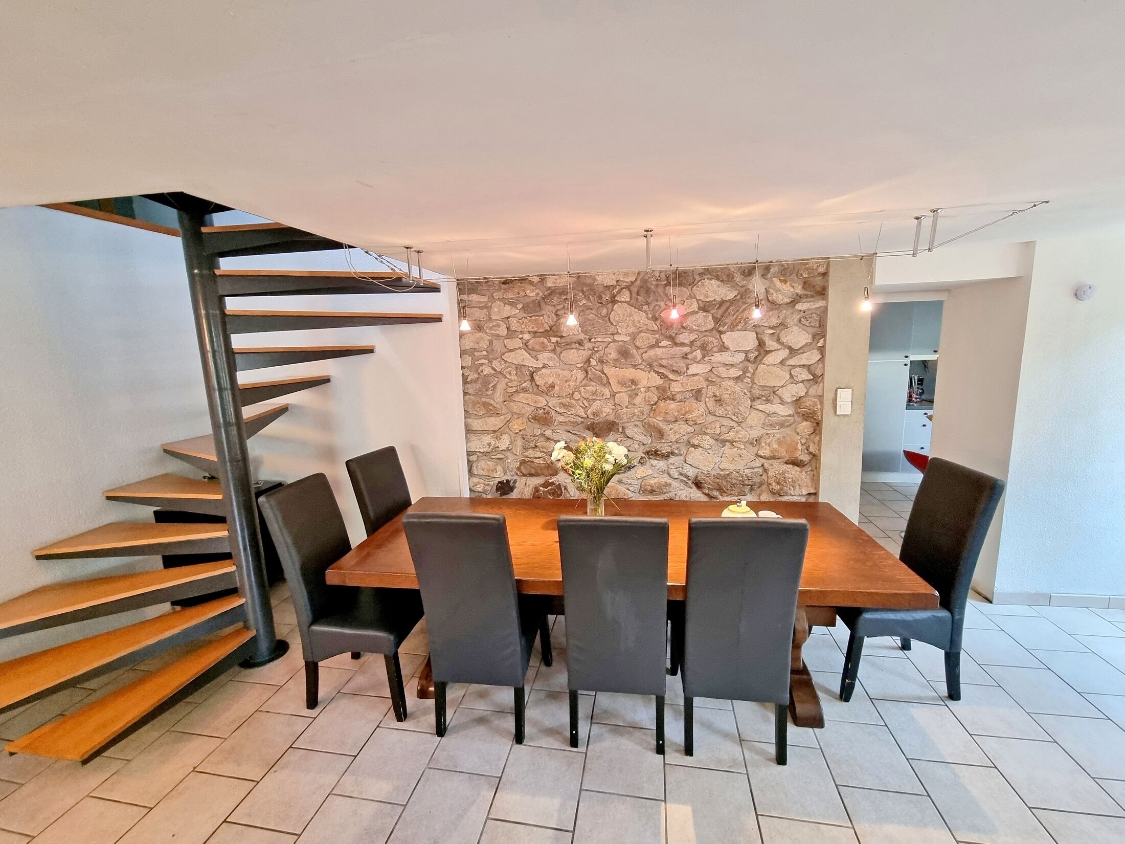 Un coin repas avec une table en bois, six chaises, un mur en pierre et un escalier en colimaçon.