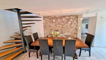 Un coin repas avec une table en bois, six chaises, un mur en pierre et un escalier en colimaçon.