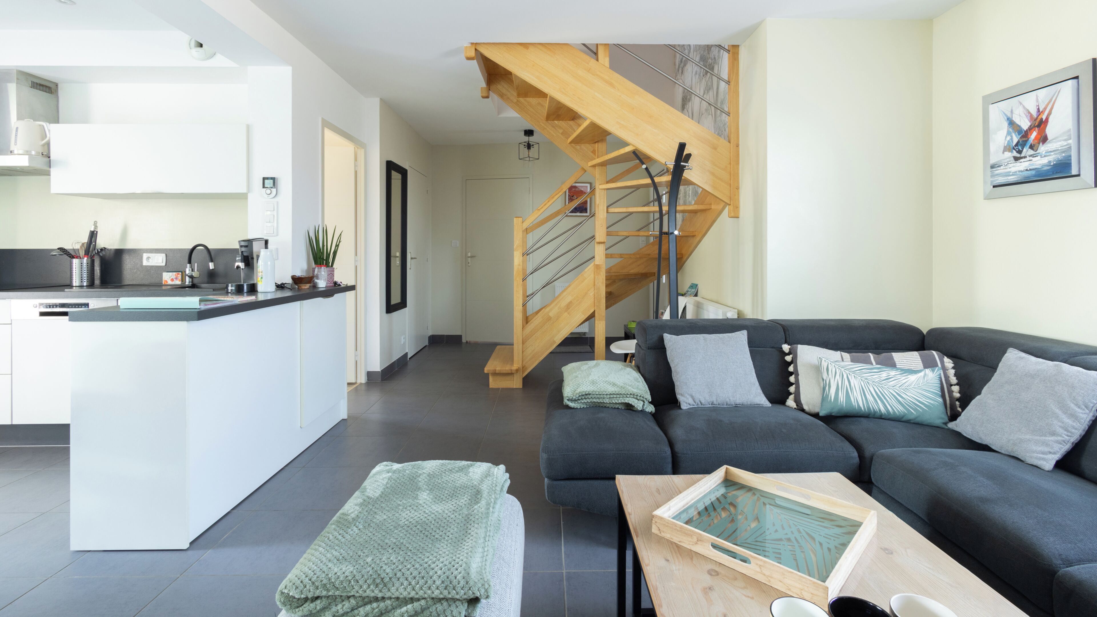 Een moderne woonruimte met een keuken, een grijze zithoek, een houten trap en een eettafel.