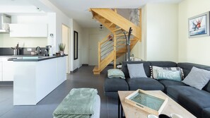 A modern living space with a kitchen, a grey sofa set, a wooden staircase, and a dining table.