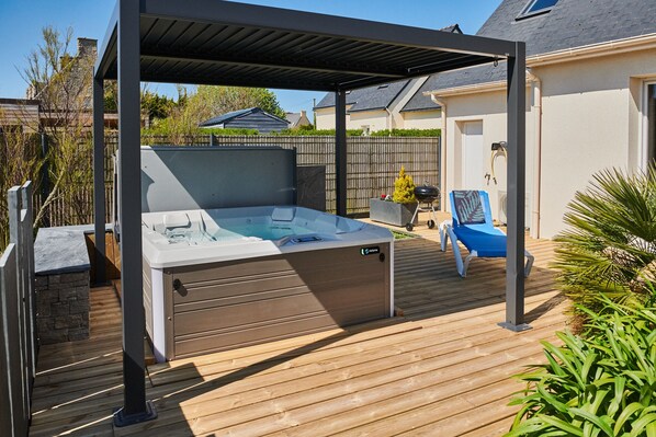 Un bain à remous sous une pergola, sur une terrasse en bois, avec vue sur une maison et une clôture en arrière-plan.