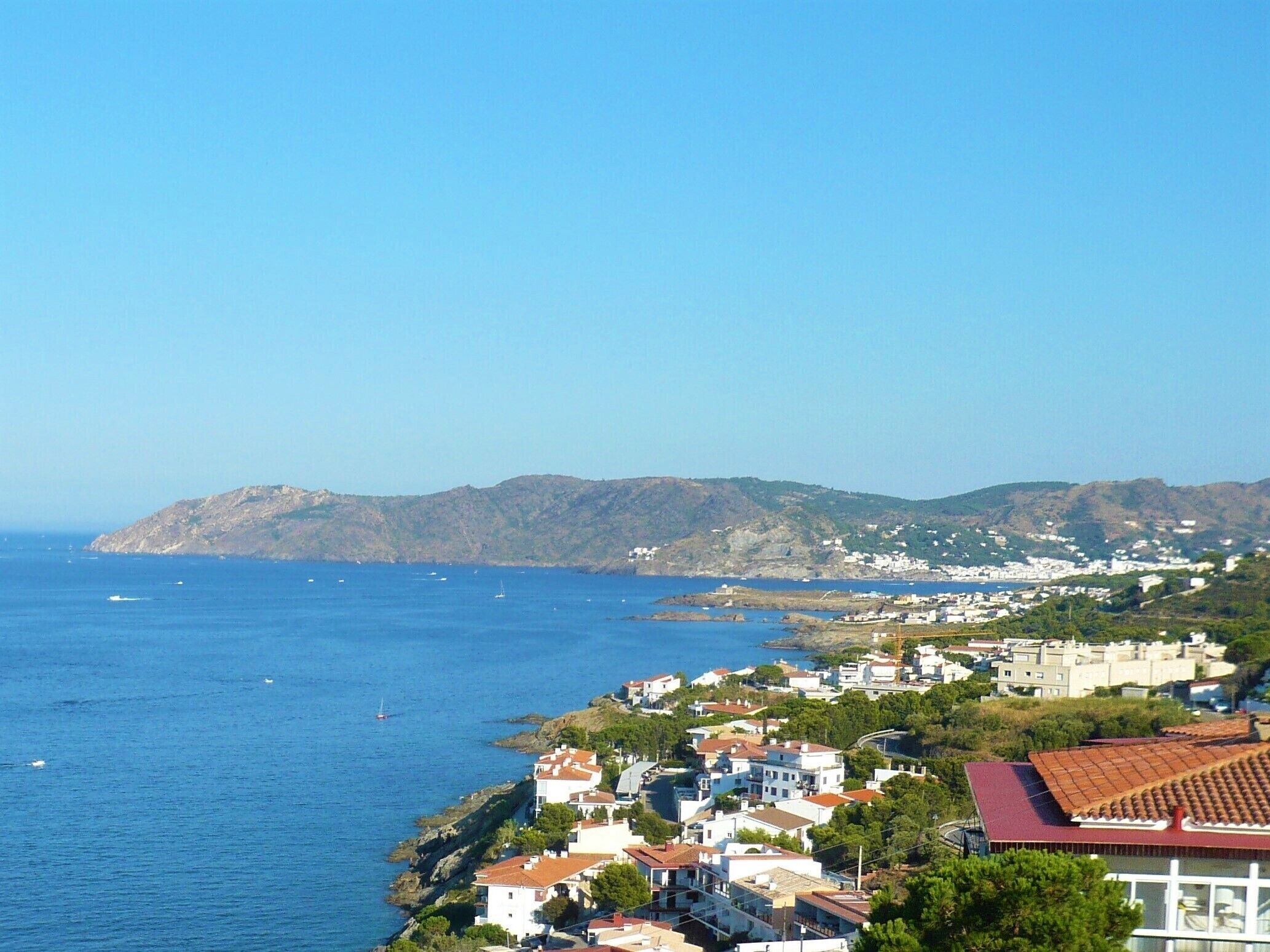 Une ville côtière avec des bâtiments qui bordent le littoral, un ciel bleu dégagé et un paysage vallonné en arrière-plan.