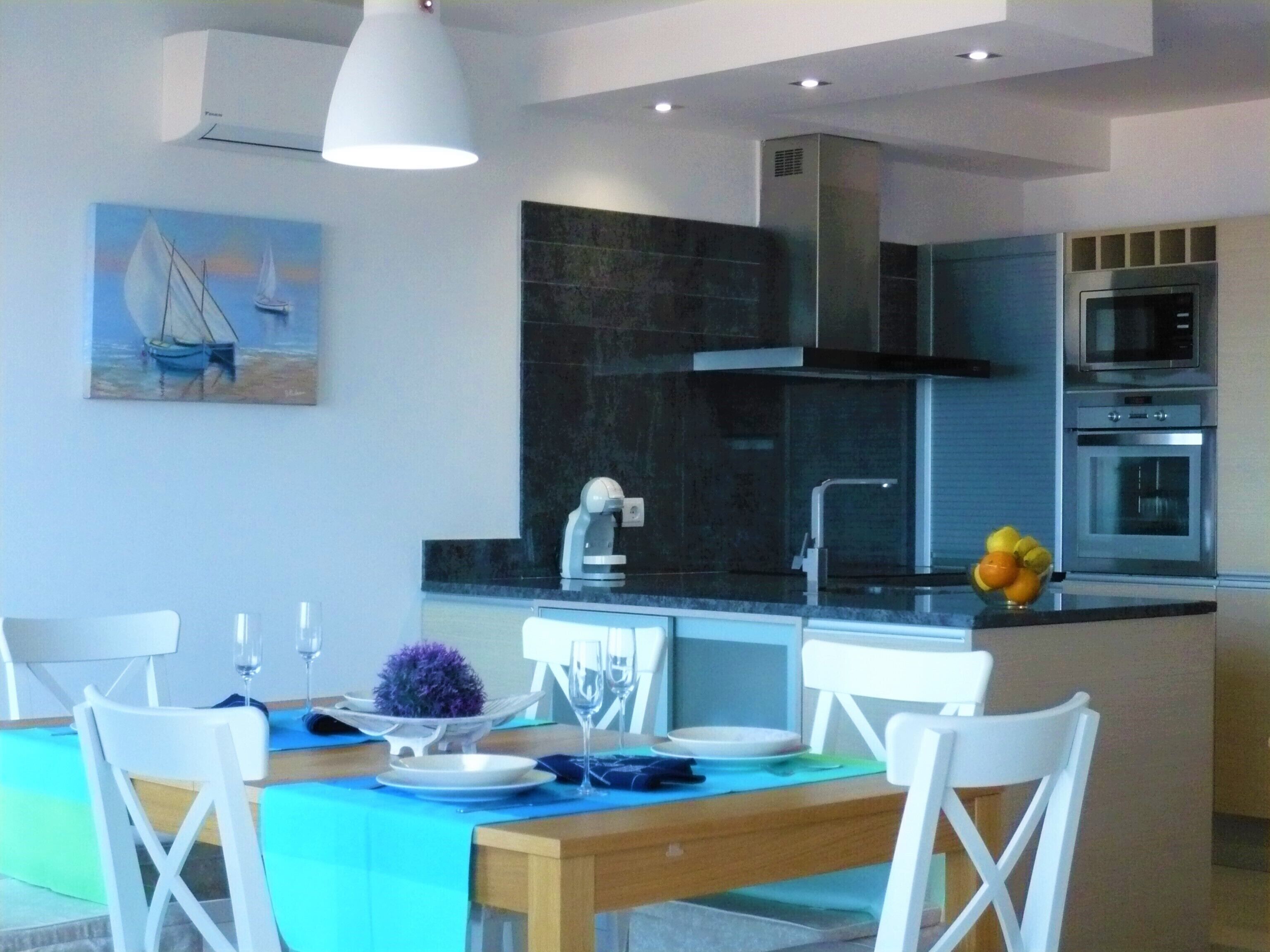 Een moderne keuken met een centraal eiland, een eetgedeelte met witte stoelen en een schilderij aan de muur.