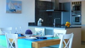 A modern kitchen with a central island, a dining area with white chairs, and a painting on the wall. - Superb new apartment, luxury, stunning sea views (Llançà)