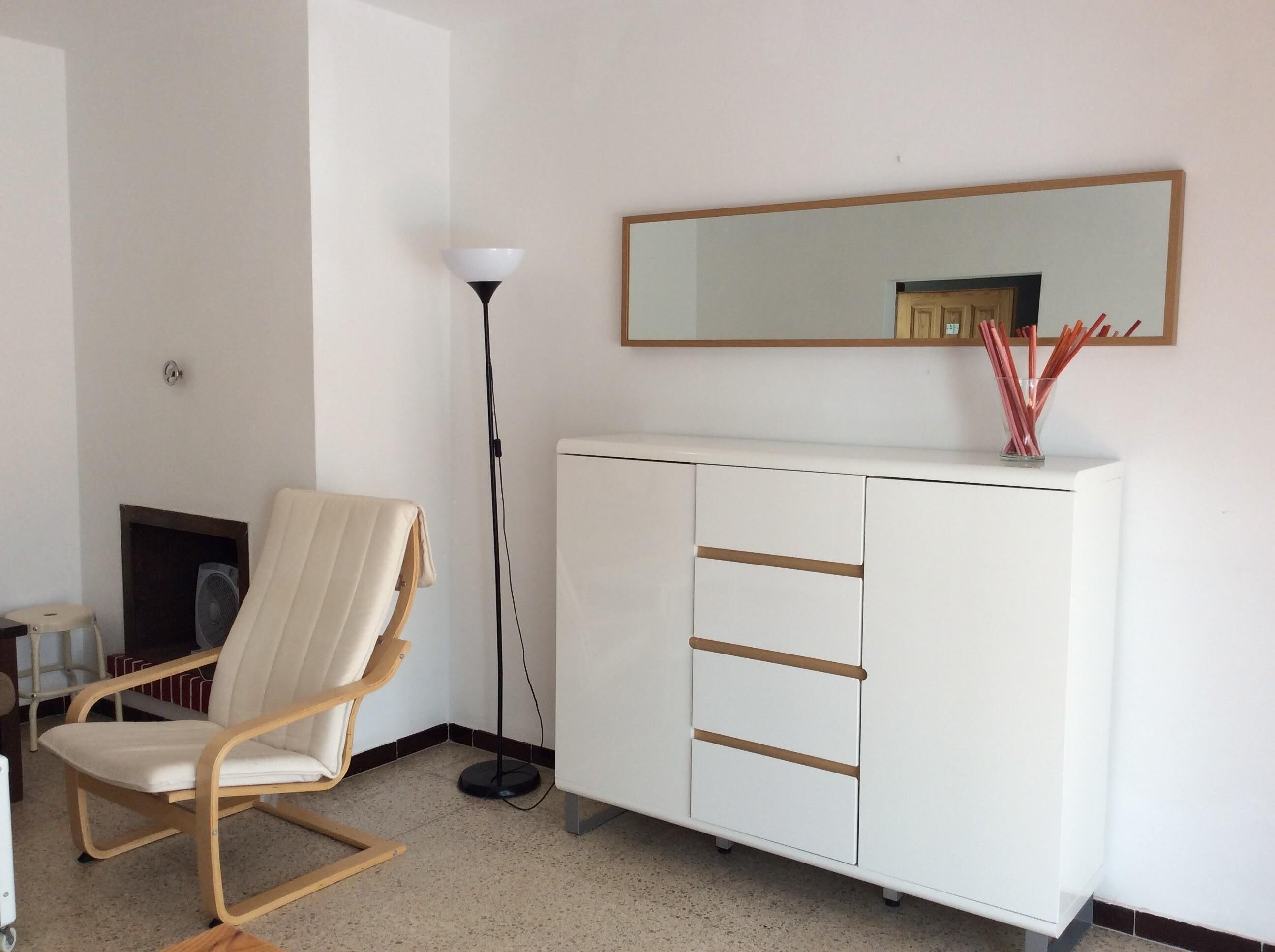 Een kamer met een wit kastje, een stoel, een vloerlamp en een spiegel aan de muur.