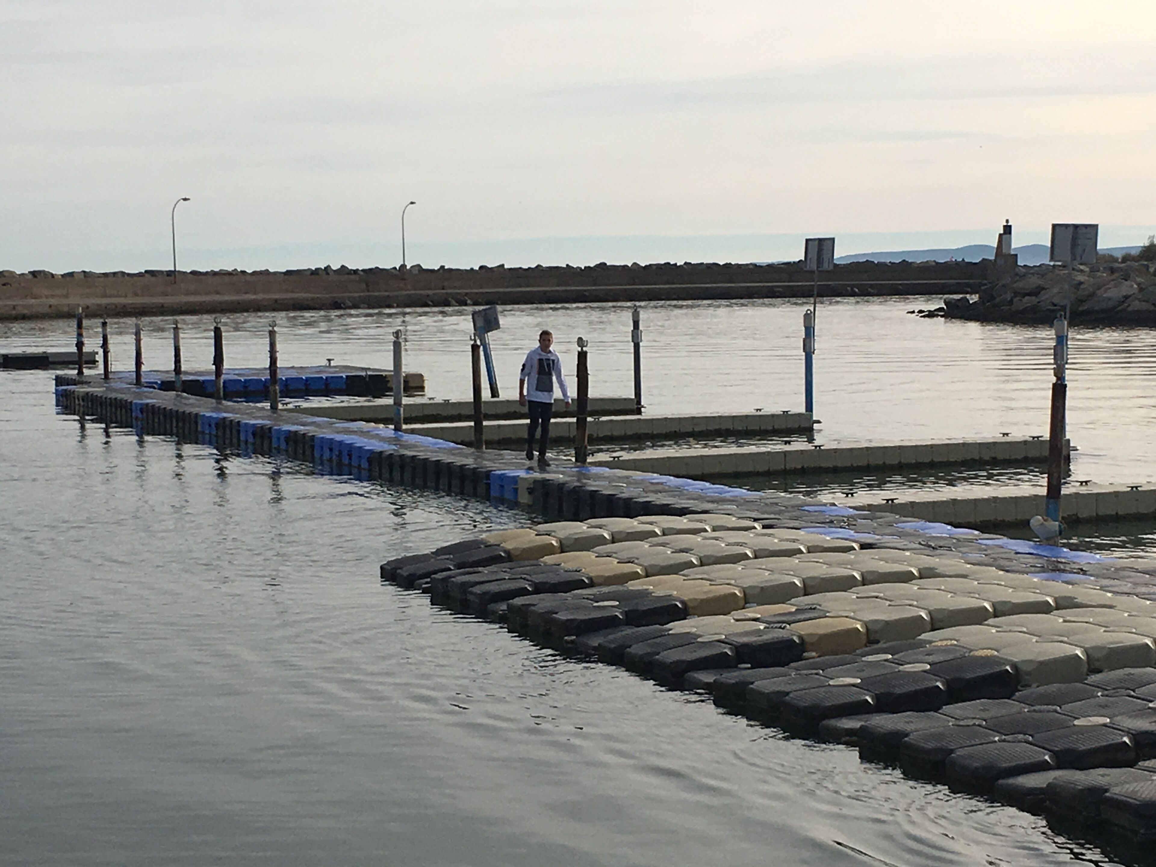 A pier with hexagonal stepping stones leading into calm water, a person standing at the end, and a distant shoreline with a lighthouse.