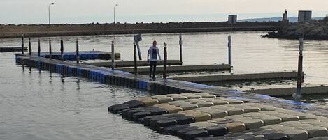 A pier with hexagonal stepping stones leading into calm water, a person standing at the end, and a distant shoreline with a lighthouse.