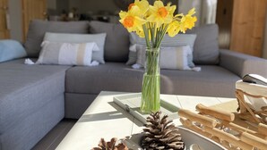 Een woonkamer met een grijze bank, een glazen vaas met gele bloemen, en een salontafel met dennenappels en houten takken.