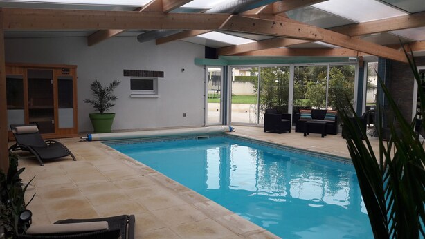 Ein Innenpool mit gefliestem Boden, Holzbalkendecke und einer Glastür, die nach draußen führt. In der Nähe des Pools stehen Liegestühle und ein Kübel mit einer Pflanze.
