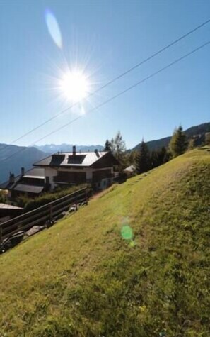 Une maison avec un toit en pente, entourée d’une colline verdoyante et avec des montagnes en arrière-plan.