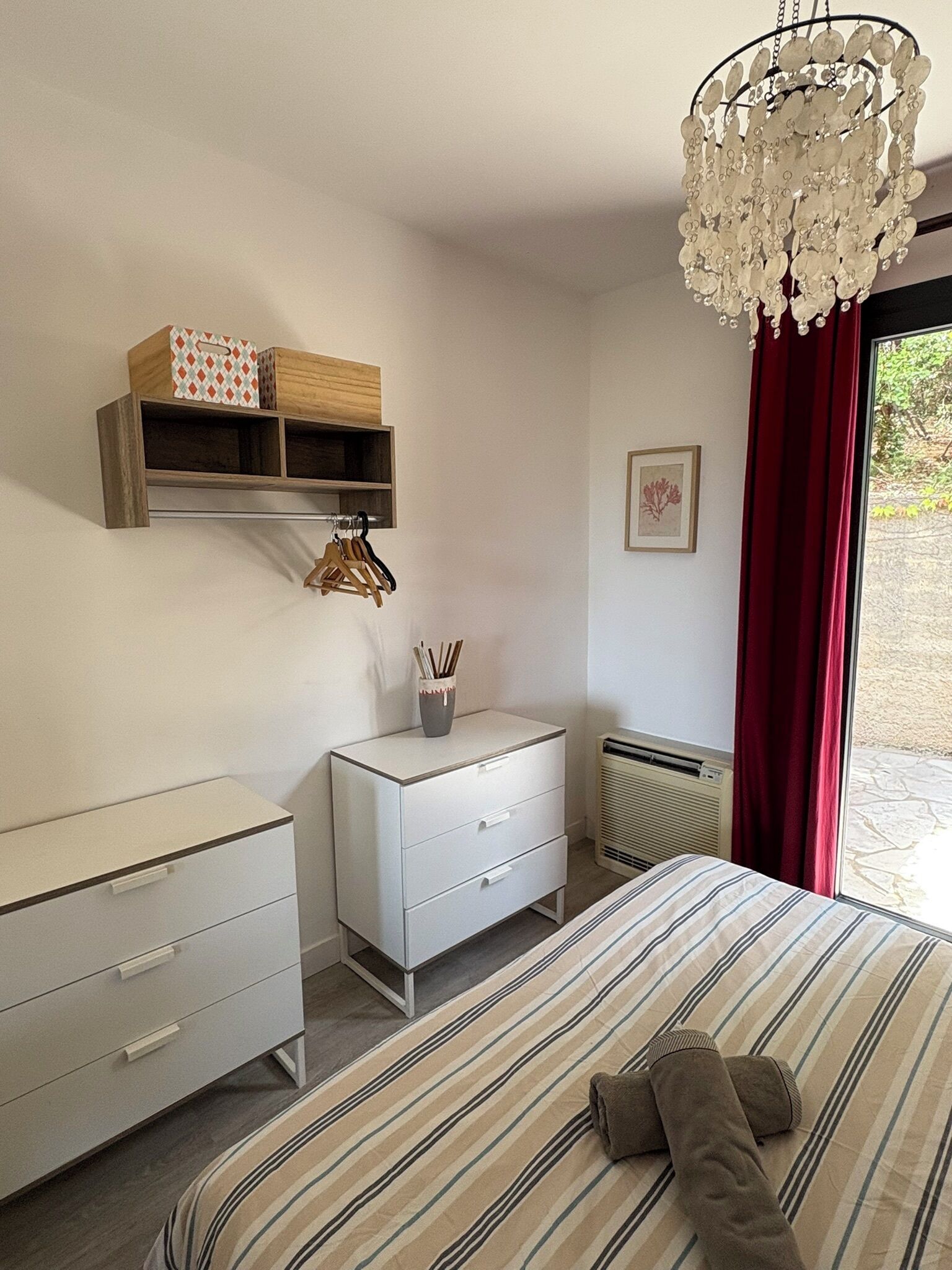 A bedroom with a bed, a bedside table, a dresser, a hanging light fixture, and a window with a view.