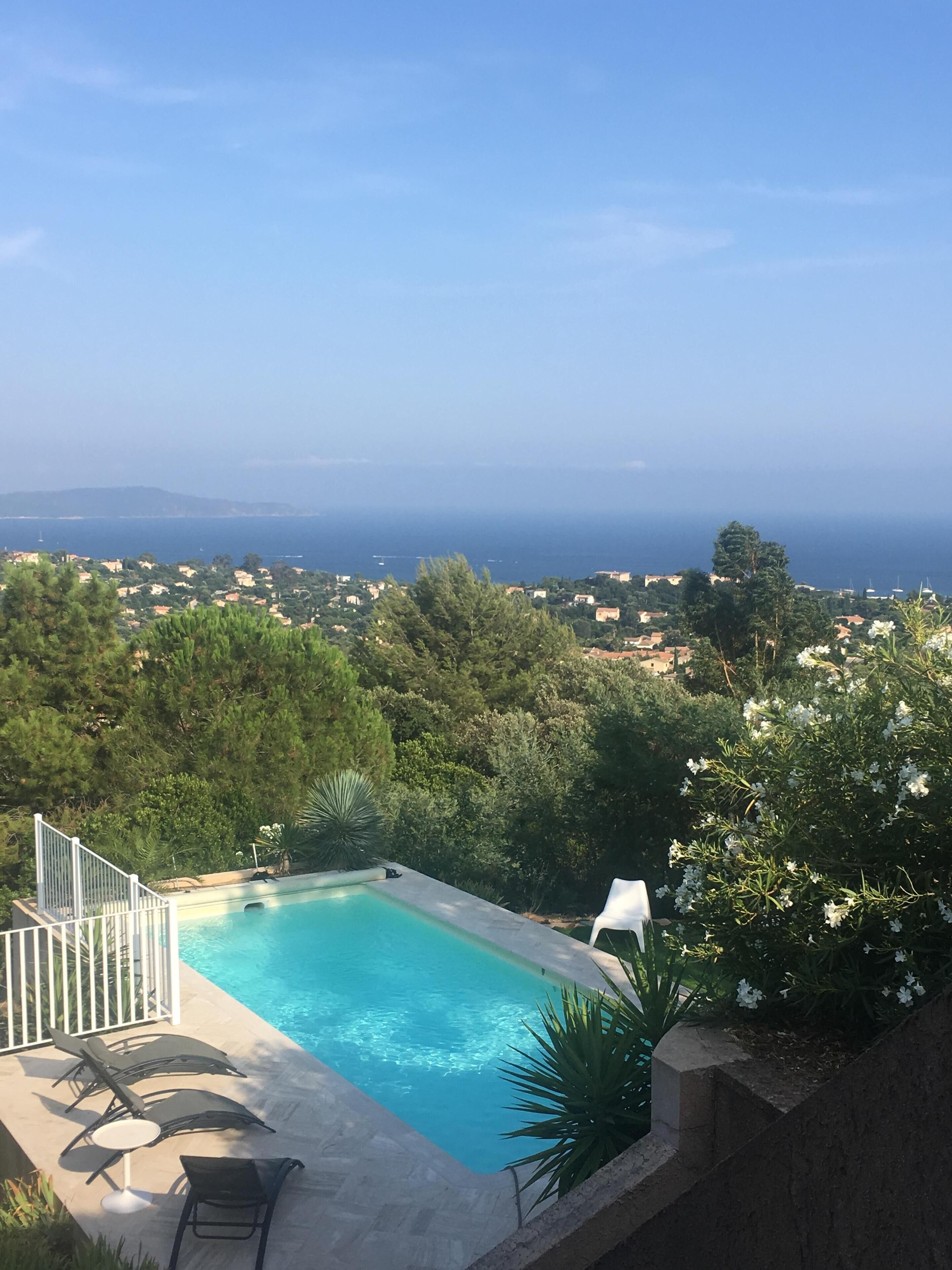 Une piscine entourée de chaises longues, bordée de verdure et offrant une vue sur une ville côtière.