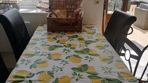 Un coin repas avec une table recouverte d’une nappe à motifs de citron, un panier en osier contenant des aliments et une vue sur la verdure à travers de grandes fenêtres.