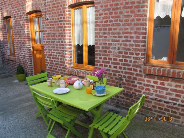 A green table set with breakfast items, a potted plant, and a window with a view of a brick building. - "La Maison", heart of the village, 50 meters from the sea (Étretat)