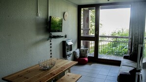 A small, cozy room with a wooden table, a television mounted on the wall, a red cushion, and a sliding glass door leading to a balcony with a view of greenery. - Apartment at the foot of the Arc 2000 slopes (BOURG SAINT MAURICE)