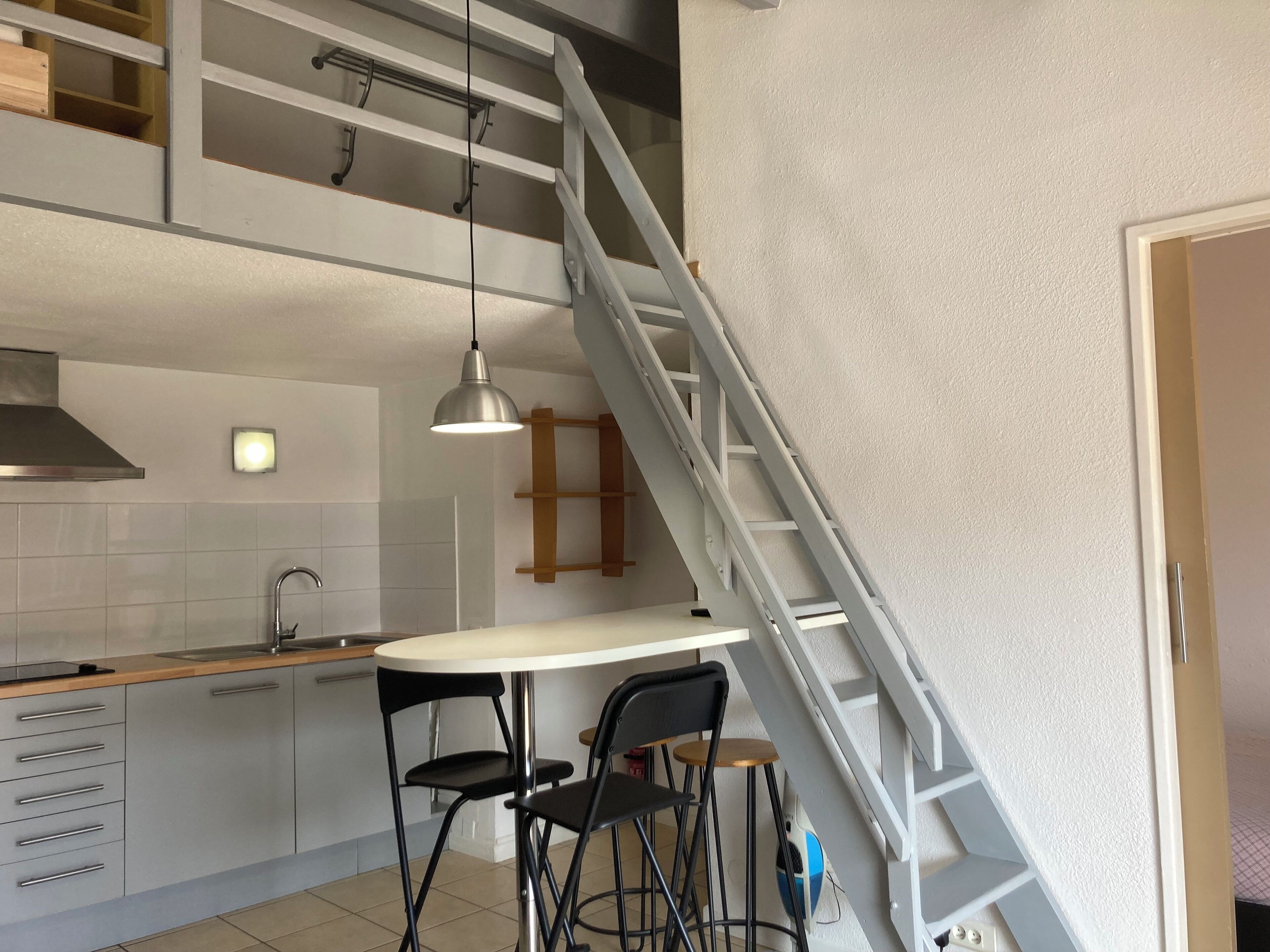 A kitchen with a white countertop, two black chairs, a stainless steel sink, and a grey staircase.