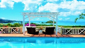 A poolside area with a pergola, outdoor seating, and a view of the ocean.