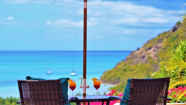 A table with two wicker chairs and an umbrella, overlooking a beach with clear blue water and boats.