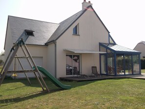 A house with a sloped roof, a covered patio area with chairs, and a green slide. - Villa, 300 m away, seaside, Agon-Coutainville, 7/8 people (Agon-Coutainville)