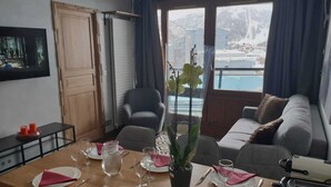 A dining area with a wooden table set for four, a black leather chair, and a sofa with cushions. A potted plant with yellow flowers is on the table. - Splendid apartment at the foot of the slopes/2 separate bedrooms/ ideal for a 4p family (Val-d'Isère)