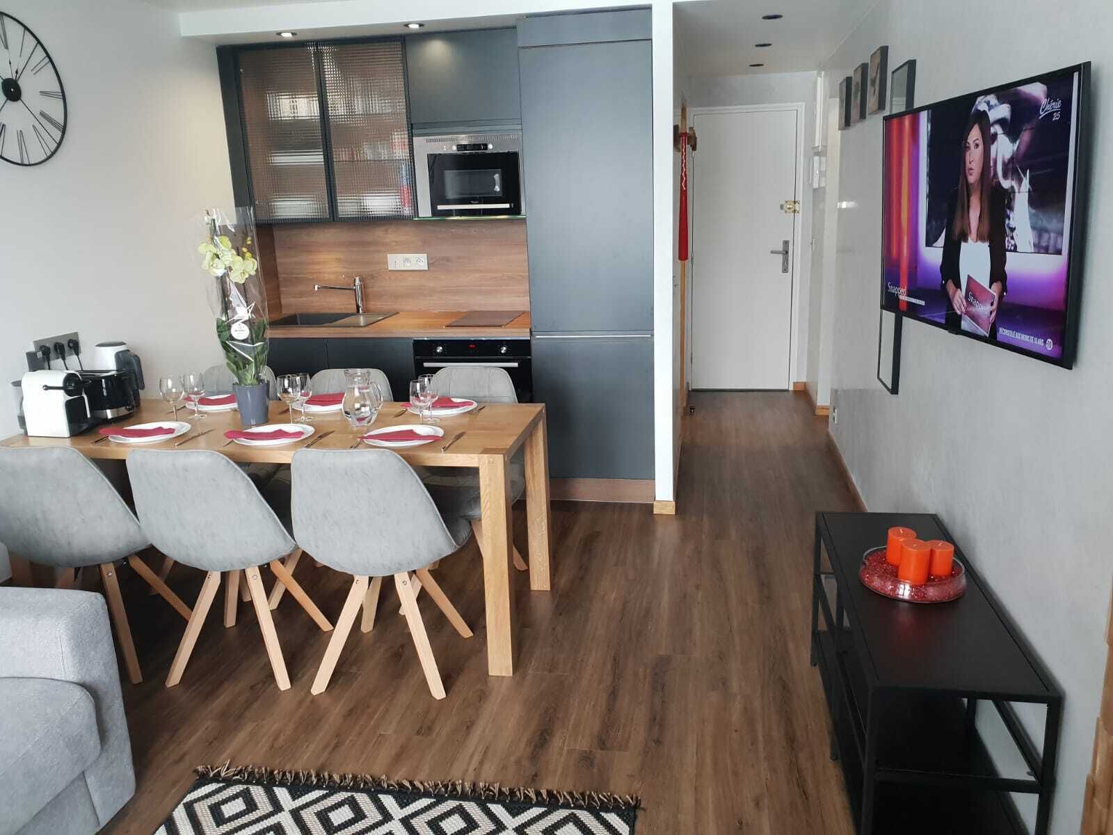 A modern kitchen with a microwave, oven, and sink. There is a dining table with chairs and a vase of flowers. A television is mounted on the wall.