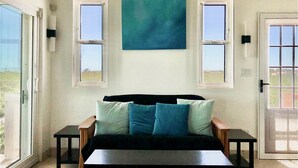 Quarto com sofá preto, duas mesinhas laterais e uma grande pintura abstrata na parede. Há duas janelas com molduras brancas e uma porta com mosquiteiro.
