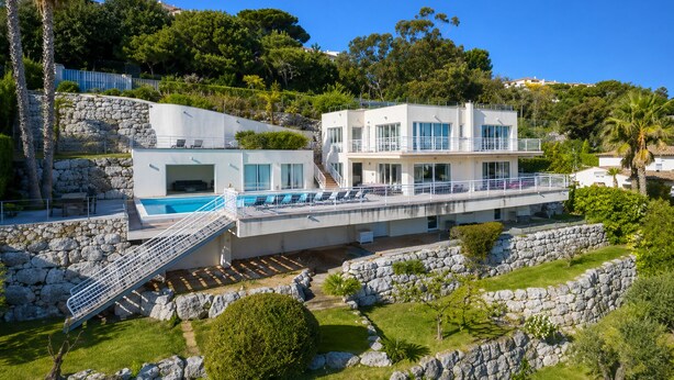 A modern two-story building with a pool, stone walls, and lush greenery.