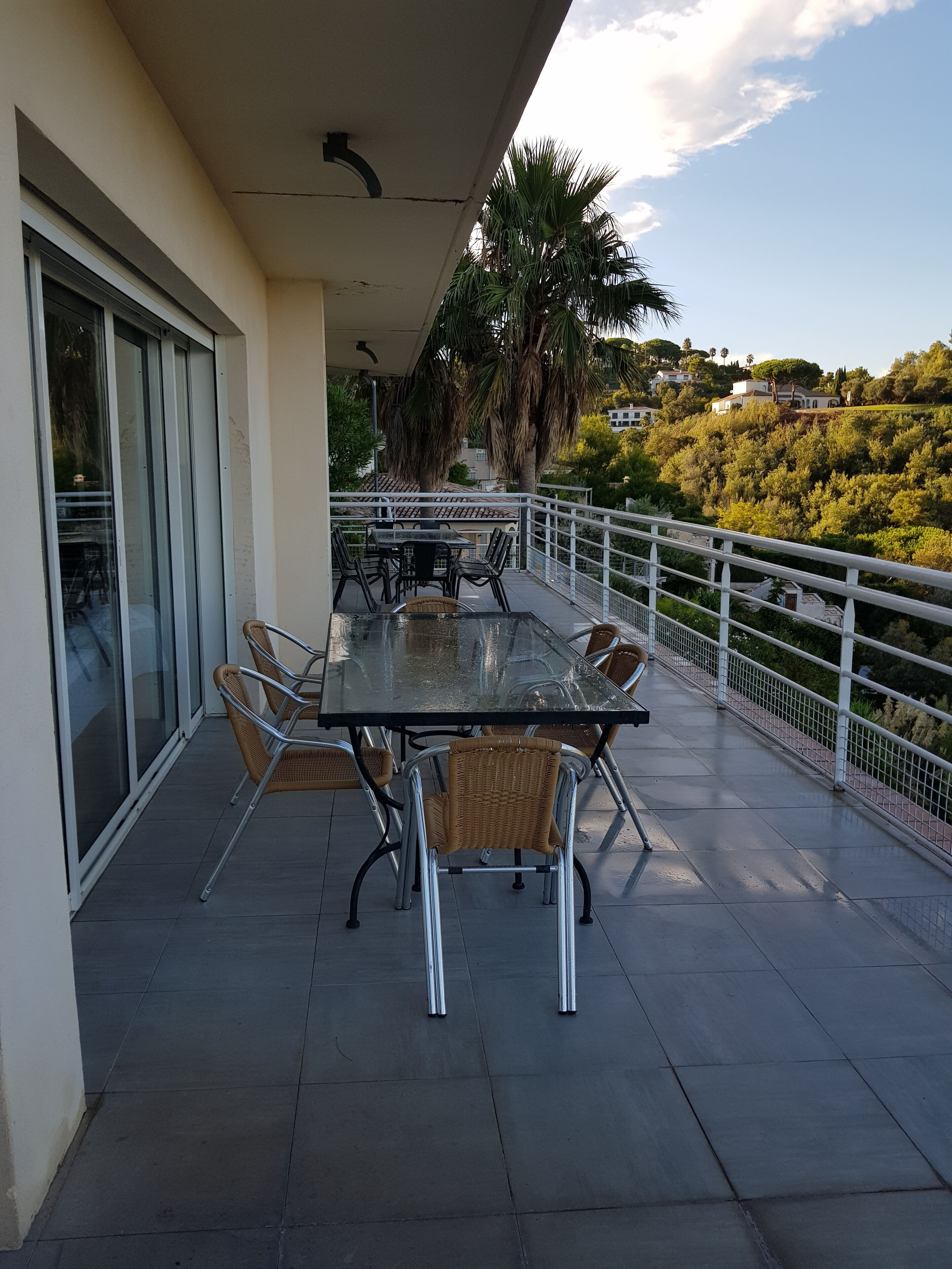 Un espace extérieur abrité, aménagé pour les repas, avec une table en verre et des chaises en osier, offrant une vue sur une colline parsemée d’arbres et de bâtiments.