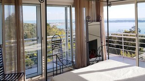 A bedroom with a large bed, a chair, and a view of a city and water through floor-to-ceiling windows.
