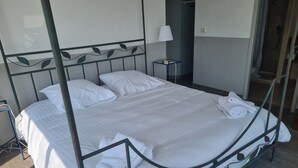 A neatly made bed with white linens, a bedside table with a lamp, and a shower area visible in the background.