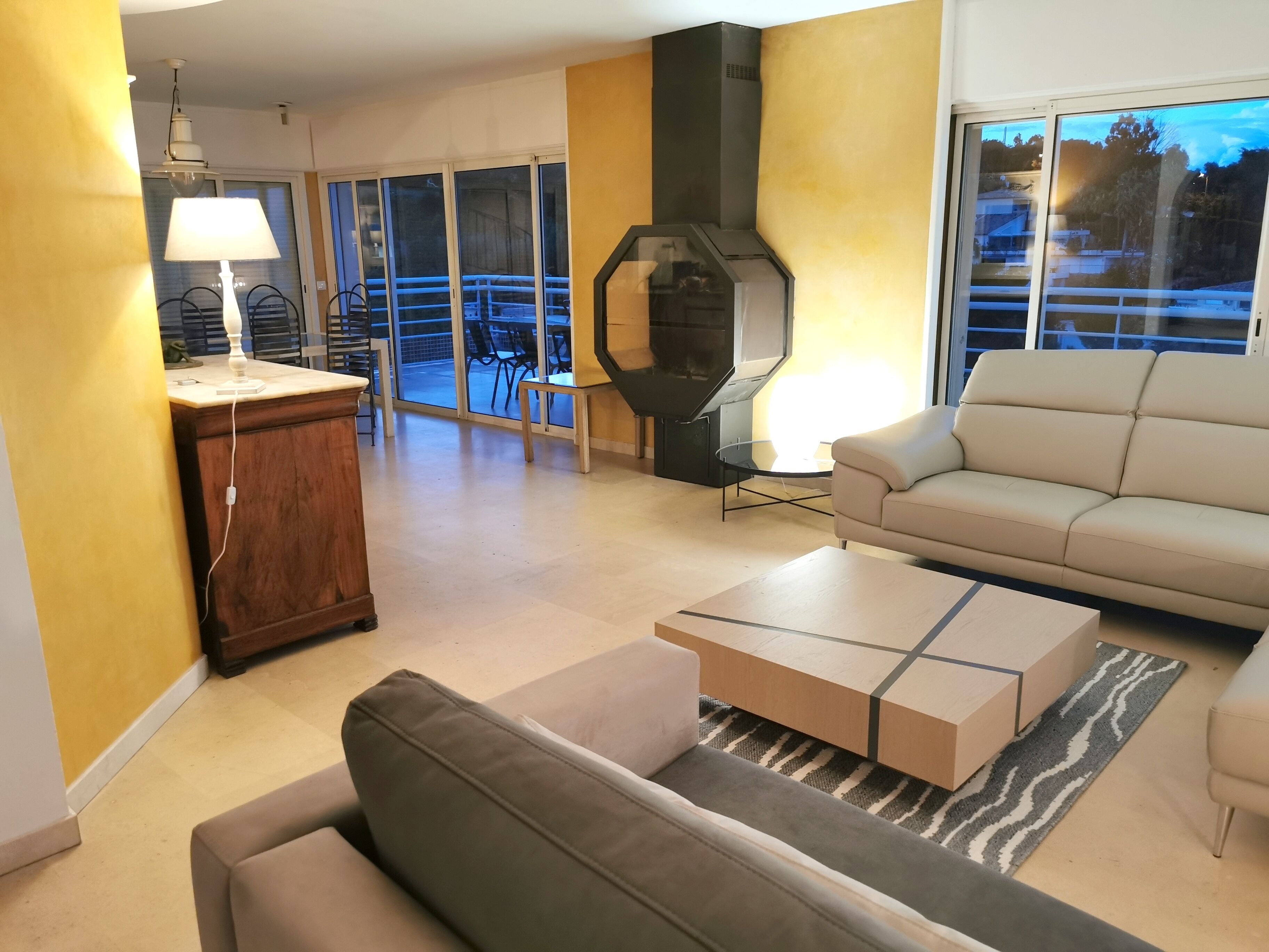 A living room with a sofa, a coffee table, a lamp, a fireplace, and a view through large windows.
