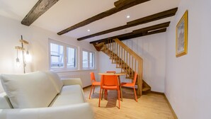 Une pièce avec un escalier en bois, des murs blancs et un plancher en bois. On y trouve un canapé blanc et des fauteuils orange disposés autour d’une petite table.