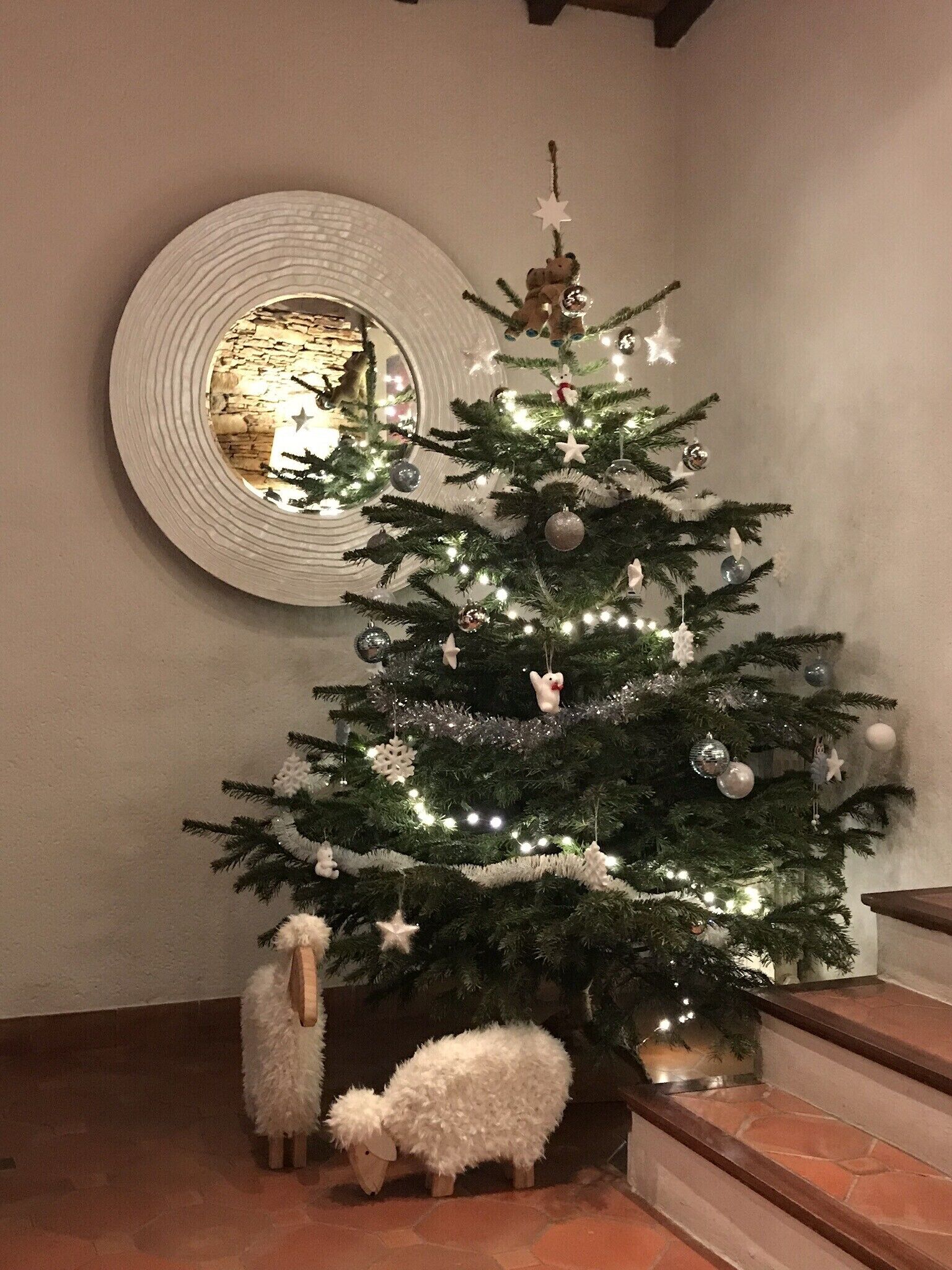 Een kerstboom versierd met lichtjes en decoraties, een spiegel aan de muur en een witte schapenbeeld aan de voet.