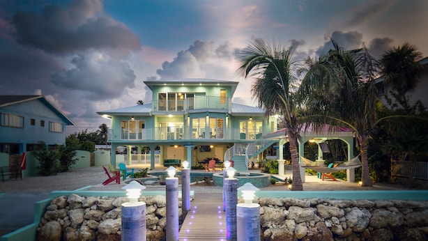 A large, two-story house with a wooden deck leading to a pool area, surrounded by palm trees and a stone wall. - Paradise Bay - 4 Bedrooms [with 2 Master Suites] & 4 Bathrooms - sleeps 10 (Marathon)