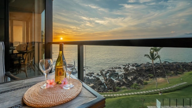 Un balcon avec vue sur l’océan au coucher du soleil, une bouteille de vin et deux verres posés sur un set de table en fibres tressées.