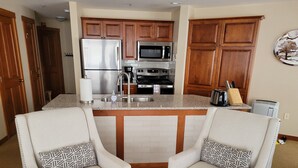 A modern kitchen with wooden cabinets, stainless steel appliances, and a central island. There are two white armchairs with patterned cushions.