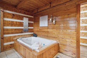Interior de una cabaña de madera con una bañera de madera grande, paredes de madera y piso de baldosas.
