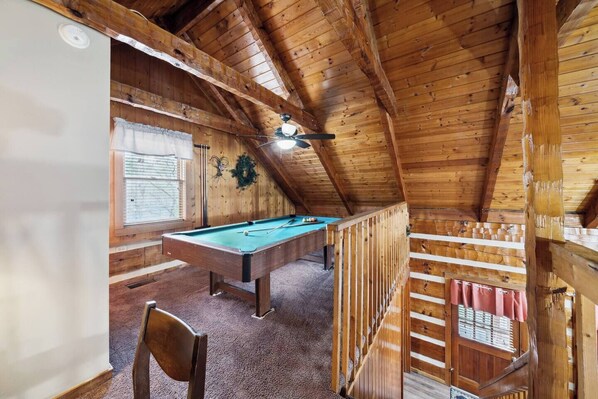 Een houten interieur met een pooltafel, een plafondventilator en een raam met zonwering.