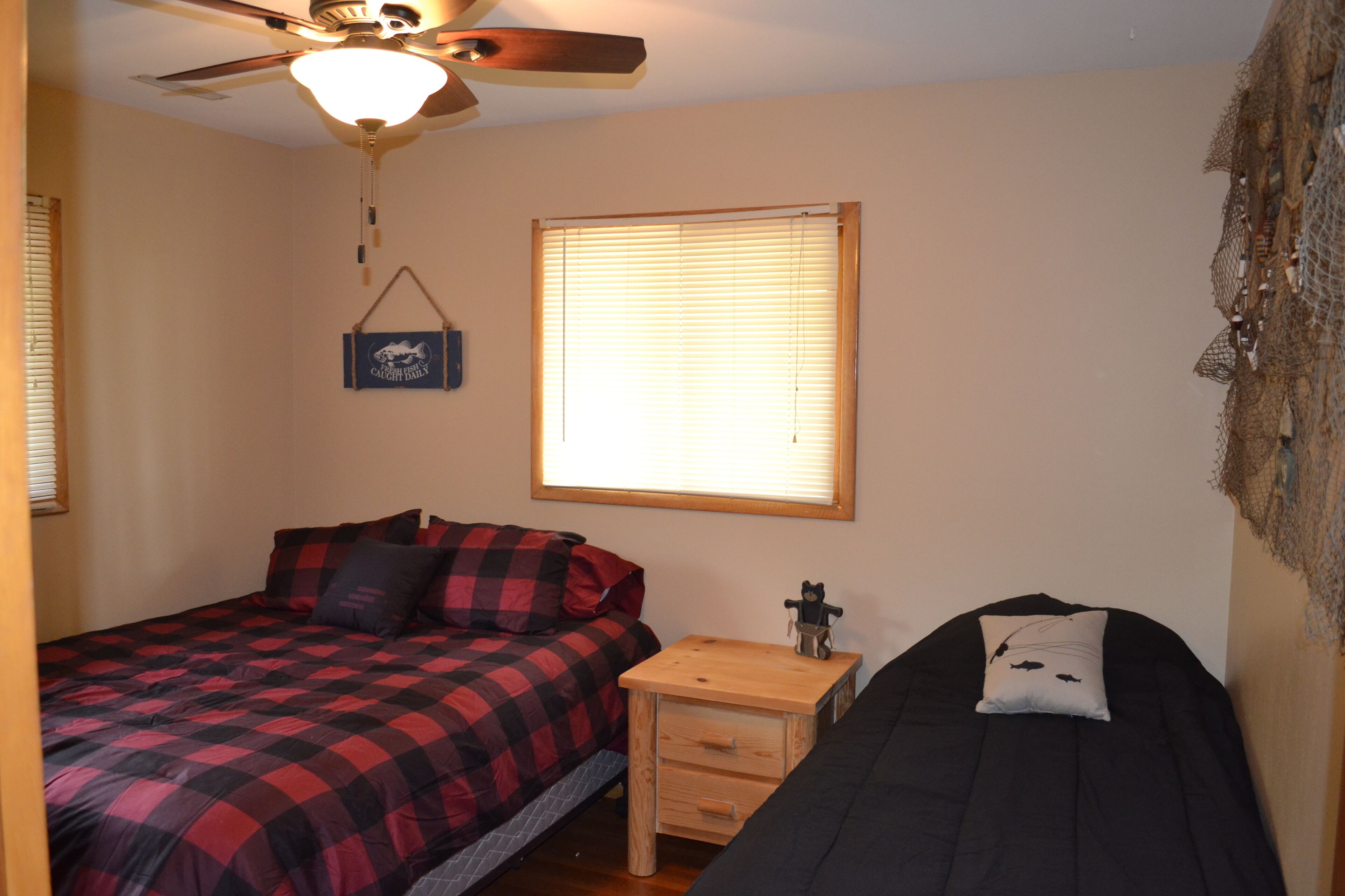 A bedroom with a bed, a nightstand, a ceiling fan, a window with blinds, and a wall-mounted shelf.