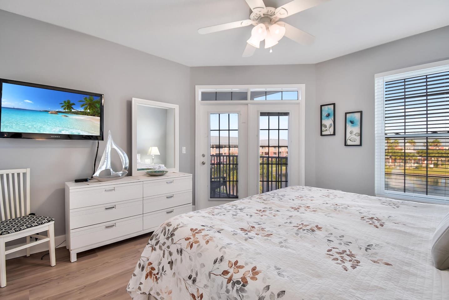 A bedroom with a bed, a dresser, a television, a ceiling fan, and a balcony with a view.