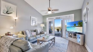 A modern living room with a grey sofa, a coffee table, and a television displaying a beach scene. - Atlantis 302D - Steps to the Ocean (St Augustine Beach)